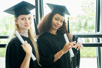 Happy Diverse Graduates Holding Diplomas, Two Young Women in Graduation Gowns and Caps, Smiling...