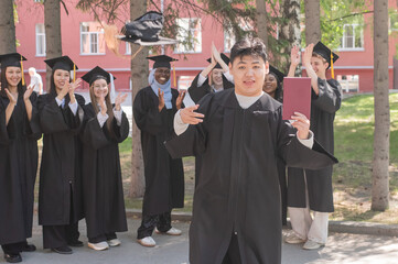 Young Asian man holds diploma while classmates applaud her. 