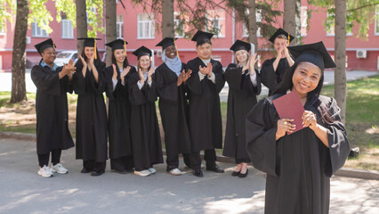 Young African woman holds diploma while classmates applaud her. 