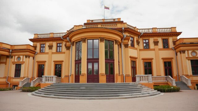 Historic yellow palace with red windows and Lithuanian national flag, viewed from front steps on a cloudy day. Elegant architecture and heritage building exterior, it's the historic Traku Vokes Manor.