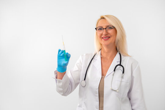 Mature blonde doctor in white uniform, gloves, and stethoscope showing a syringe prepared for injection. Isolated on white background