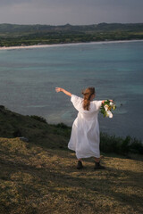 A young woman in a white dress with a bridal bouquet in her hands walks through the hills against...