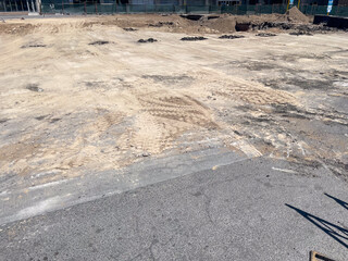 A large, cleared construction site features uneven, light brown earth with tire tracks. Surrounding...