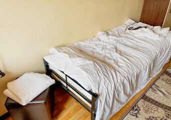 A neatly unmade bed features crumpled white linens, a brown leather ottoman nearby, and a small...