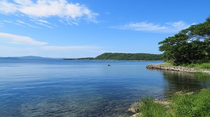 Fototapeta premium Serene lakeside view on a sunny day, clear blue water, lush green shoreline, and distant hills under a partly cloudy sky