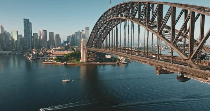 Australia, Sydney: Sydney Harbour Bridge and city skyline, sailboat glides under sunny morning blue sky. Travel destination, modern cityscape architecture in background. Aerial view drone footage
