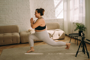 A young curly-haired plus-size woman practices yoga at home, online broadcast.Healthy lifestyle.