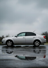 Gray sedan parked on a wet street on a cloudy day. Side view of the vehicle reflecting in a puddle, showcasing the car's profile.