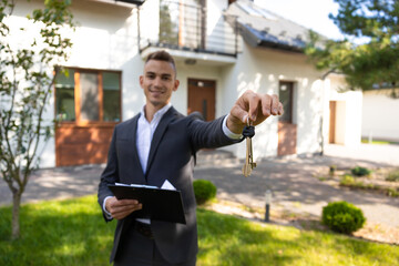 Real estate agent presenting house keys and documents on clipboard in front of property for sale