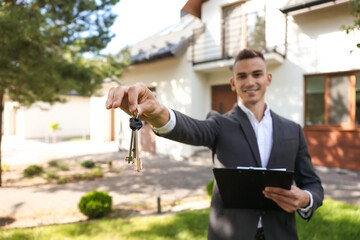 Real estate agent presenting house keys and documents on clipboard in front of property for sale