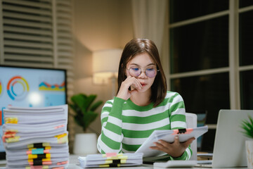 A stressed woman works late at a cluttered desk, surrounded by papers and charts, overwhelmed with...