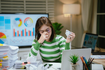 A stressed woman works late at a cluttered desk, surrounded by papers and charts, overwhelmed with...