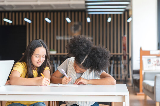 Diverse Young Girls Studying Together at School, Two Elementary Students Focused on Writing in Classroom, Kids Collaborating on Homework at a Desk