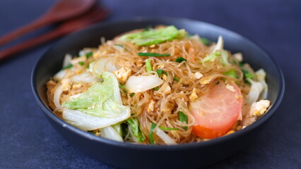 Stir-fried glass noodle with egg and vegetables in black bowl.