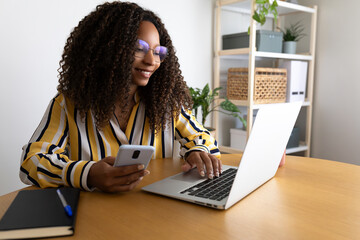 Happy African American woman using phone and laptop working at home office.