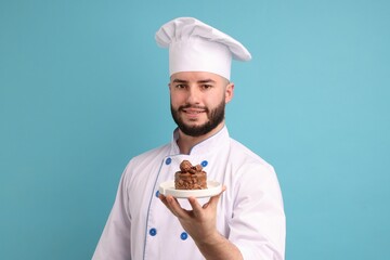 Happy confectioner in uniform holding delicious chocolate dessert on light blue background