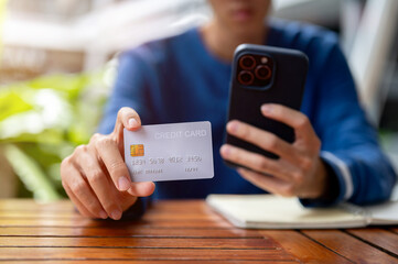 Close up of hand holding phone and man looking at his credit card on wooden table in cafe or canteen