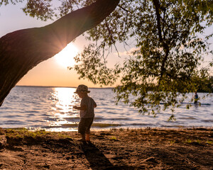 Toddler is playing by the river with joy during sunset under a tree. 
