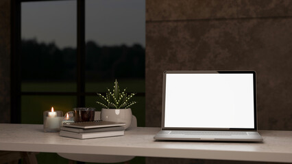 Blank screen laptop with books aside aroma candle and potted plant on a white table in dim lit room