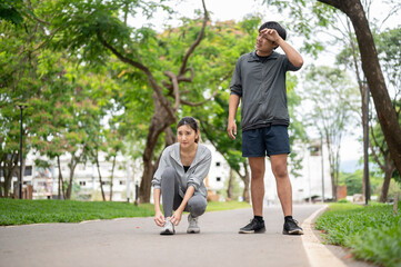 Obraz premium Asian woman in sportswear tying shoelace and a man wiping sweat after running on pavement in park.