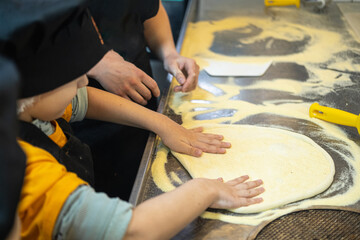 A young Asian boy with short black hair shapes dough on a floured table. Adult hands assist him.