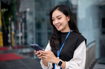 Smiling office worker or event staff holding and looking at phone while standing outside a building.