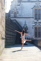 Fototapeta premium Graceful ballerina in soft pink dress dancing on pointe in sunlit alley with historic French castle in the background