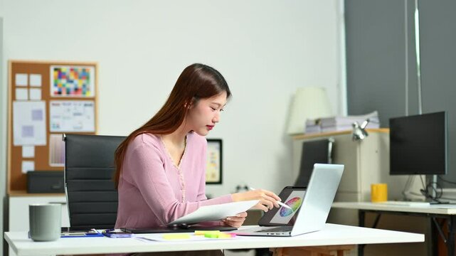 Young female graphic designer using digital tablet and laptop in modern office. Creative workspace, color palettes, and UI UX design