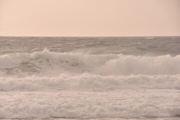 Powerful ocean waves crashing under a soft pastel-toned sky during sunset. The golden light of the setting sun casts a warm glow over the scene, creating an atmosphere that is both serene and dramatic