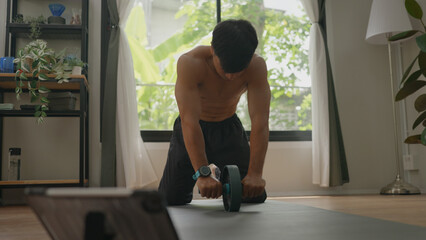 Asian man is exercising at home using an ab roller to strengthen his abdominal muscles and improve...