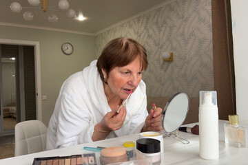 A beautiful senior woman is sitting at home and doing her make-up morning routine. Senior woman putting blush on her face