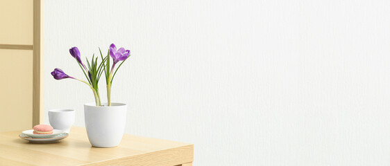 Pot with beautiful crocus flowers, cup and tasty macaroon on table near white wall