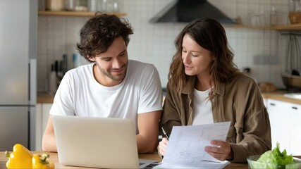 Happy couple reviewing finances together in their kitchen using a laptop. - Powered by Adobe
