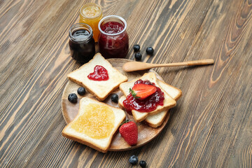 Plate with delicious toasts, jars of different jams and berries on brown wooden background
