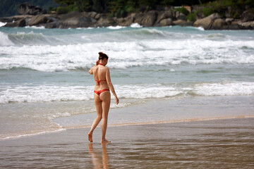 Young woman in bikini walking by sand of sea beach on breaking waves and stones background, summer...