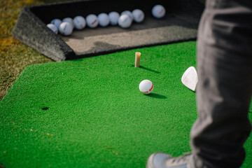 Close-up of a golfer preparing to hit a ball on a green practice mat, with multiple golf balls and a club visible, during a sunny outdoor session.
