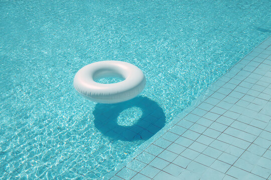 Bright turquoise swimming pool with clean white tiles and a single inflatable ring float drifting off-center, sun casting soft reflections