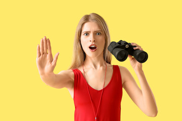 Young female lifeguard with binocular showing STOP gesture on yellow background