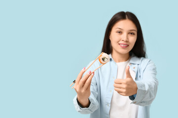 Beautiful young Asian woman holding tasty sushi roll with chopsticks and showing thumb-up on pale...