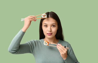 Beautiful young Asian woman holding tasty sushi rolls with chopsticks on green background