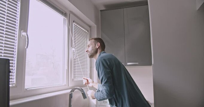 A young man opens the blinds in his kitchen, letting in the morning sunlight. He begins his daily routine at home.