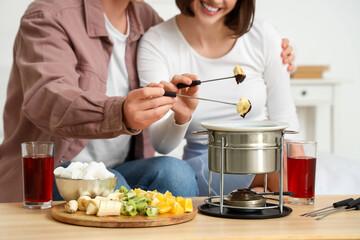 Young happy couple with glasses of wine dipping fresh bananas into chocolate fondue at home, closeup