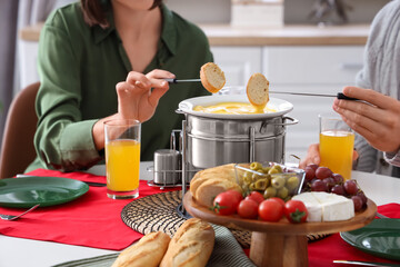 Young couple with glasses of juice dipping croutons into cheese fondue at home, closeup
