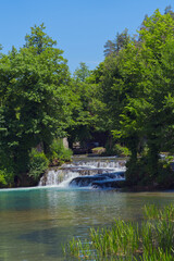 A serene natural scene featuring cascading waterfalls emptying into a clear, tranquil pool, surrounded by lush green trees under a bright blue sky.