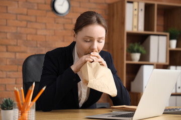 Young businesswoman with paper bag having panic attack at table in office