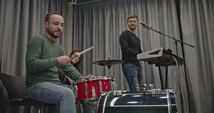 A music band practices in their studio before a concert. The drummer plays a red drum kit, the keyboard player smiles, and the guitarist strums. They are preparing their music.