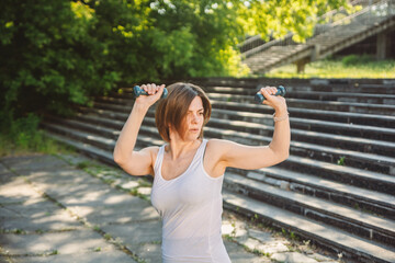 Woman exercising outdoors with dumbbells, lifting weights on stairs. Fitness training for health...