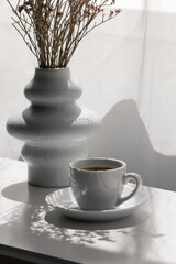 Cup of hot coffee and vase with gypsophila flowers on white table in room