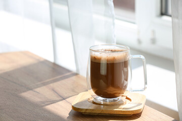 Glass cup of hot coffee on wooden table near window in room