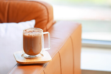 Glass cup of hot coffee on sofa near window in living room